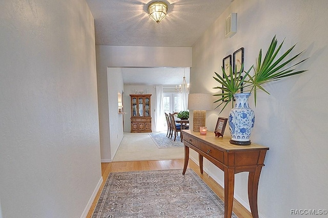 corridor featuring a textured ceiling, a notable chandelier, visible vents, baseboards, and light wood finished floors