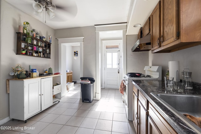 kitchen featuring light tile patterned floors, brown cabinetry, white gas stove, under cabinet range hood, and a sink