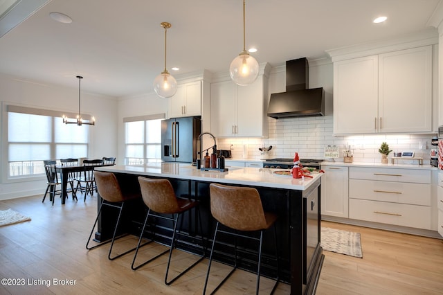 kitchen with wall chimney range hood, a kitchen island with sink, white cabinets, high end refrigerator, and light wood-type flooring