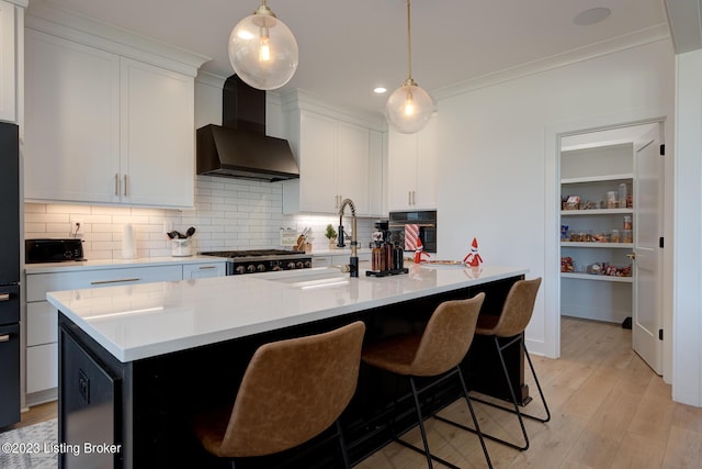 kitchen with wall chimney exhaust hood, white cabinetry, light hardwood / wood-style flooring, an island with sink, and pendant lighting