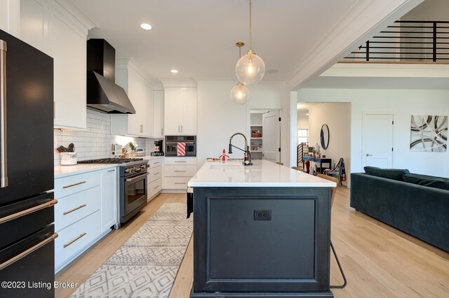 kitchen with sink, a kitchen island with sink, hanging light fixtures, high end appliances, and wall chimney exhaust hood