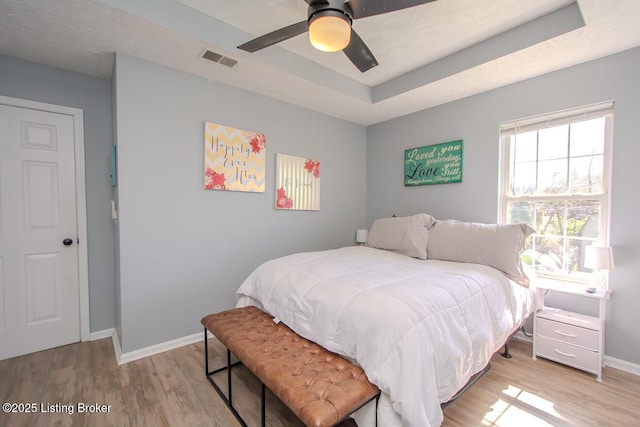 bedroom featuring light wood-type flooring, baseboards, visible vents, a tray ceiling, and ceiling fan