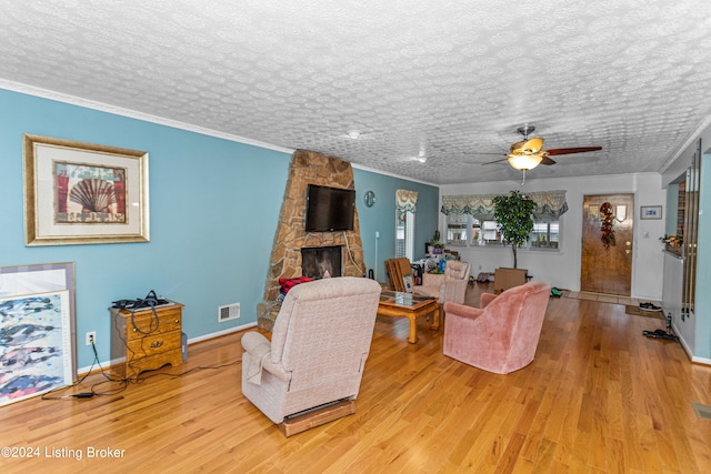 living room with light wood-type flooring, ceiling fan, a textured ceiling, a stone fireplace, and ornamental molding