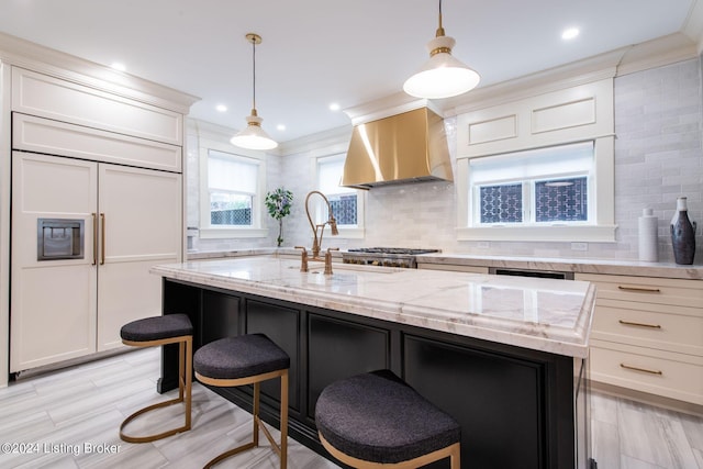 kitchen featuring wall chimney exhaust hood, hanging light fixtures, a center island with sink, and crown molding