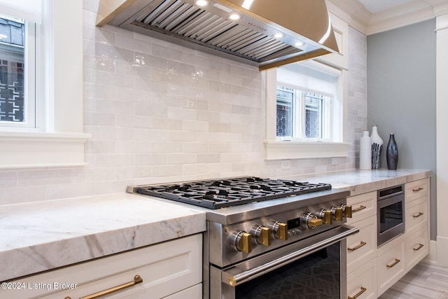 kitchen featuring stainless steel appliances, white cabinetry, light stone countertops, backsplash, and wall chimney range hood