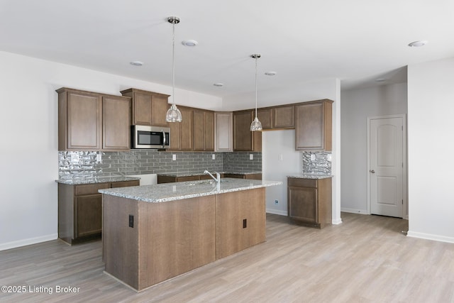 kitchen featuring a center island with sink, sink, hanging light fixtures, tasteful backsplash, and light stone counters