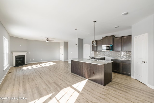 kitchen with backsplash, dark brown cabinetry, a kitchen island with sink, ceiling fan, and hanging light fixtures