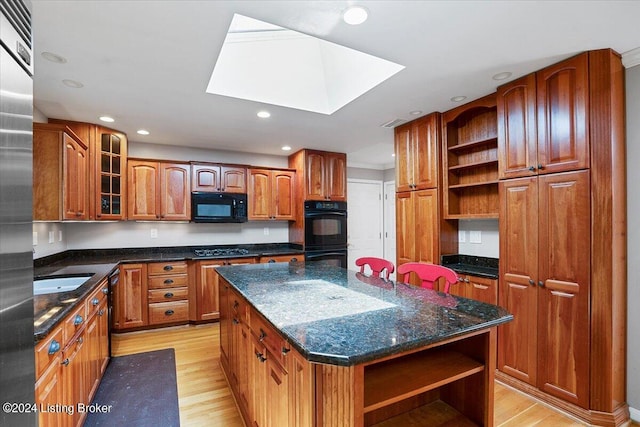 kitchen with a skylight, black appliances, light hardwood / wood-style flooring, dark stone countertops, and a center island