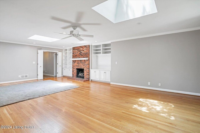 unfurnished living room featuring a skylight, a brick fireplace, crown molding, built in features, and light hardwood / wood-style flooring