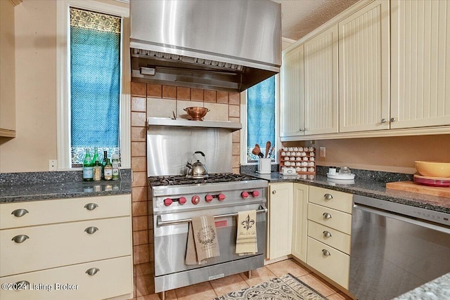 kitchen featuring stainless steel appliances, wall chimney range hood, dark stone counters, and cream cabinetry