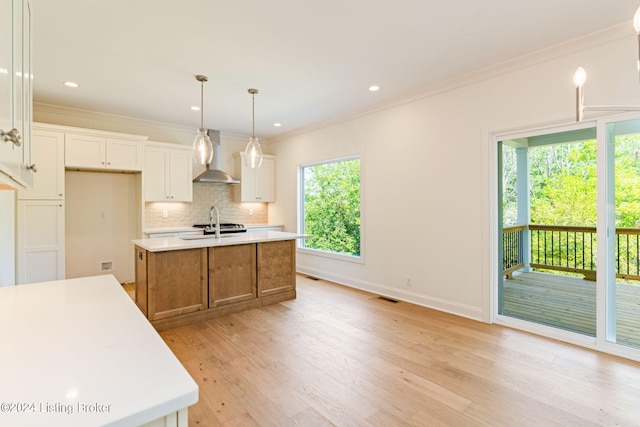 kitchen with decorative light fixtures, wall chimney range hood, white cabinets, and a kitchen island with sink