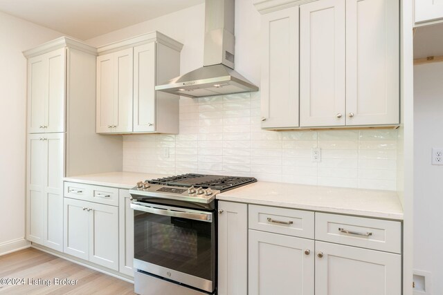 kitchen featuring wall chimney exhaust hood, light hardwood / wood-style flooring, stainless steel gas range, decorative backsplash, and white cabinetry