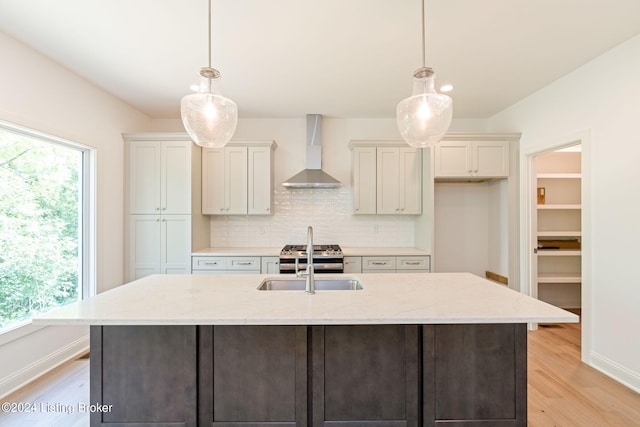 kitchen featuring wall chimney range hood, light stone countertops, light wood-type flooring, and an island with sink