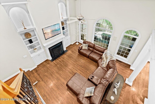 living room with built in shelves, a fireplace with flush hearth, a ceiling fan, wood finished floors, and baseboards