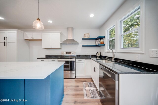 kitchen featuring stainless steel appliances, wall chimney range hood, pendant lighting, dark stone countertops, and white cabinetry