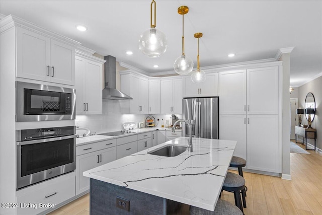 kitchen featuring pendant lighting, light hardwood / wood-style floors, wall chimney range hood, a center island with sink, and appliances with stainless steel finishes