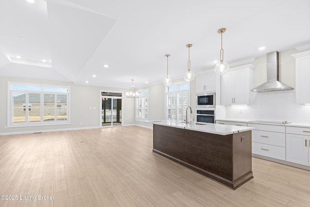 kitchen featuring wall chimney range hood, white cabinetry, an island with sink, built in microwave, and stainless steel oven