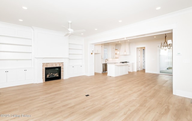 unfurnished living room featuring a brick fireplace, built in shelves, ceiling fan with notable chandelier, and ornamental molding