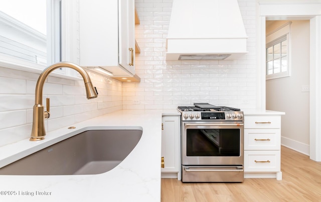 kitchen featuring sink, stainless steel gas range oven, custom range hood, light hardwood / wood-style floors, and white cabinets