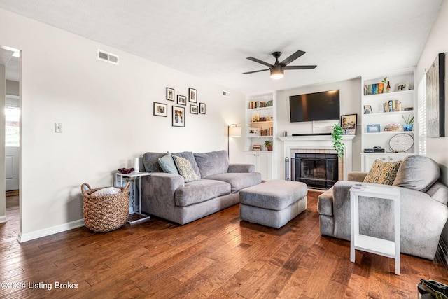 living room with a textured ceiling, hardwood / wood-style flooring, built in shelves, a tiled fireplace, and ceiling fan