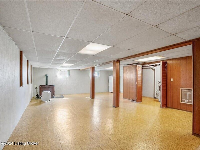 basement featuring wood walls, a drop ceiling, a wood stove, heating unit, and white refrigerator