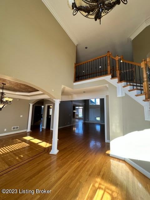 unfurnished living room featuring a high ceiling, ornate columns, wood-type flooring, crown molding, and a notable chandelier