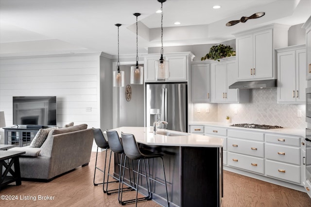 kitchen with light wood-type flooring, hanging light fixtures, white cabinetry, stainless steel appliances, and a kitchen island with sink