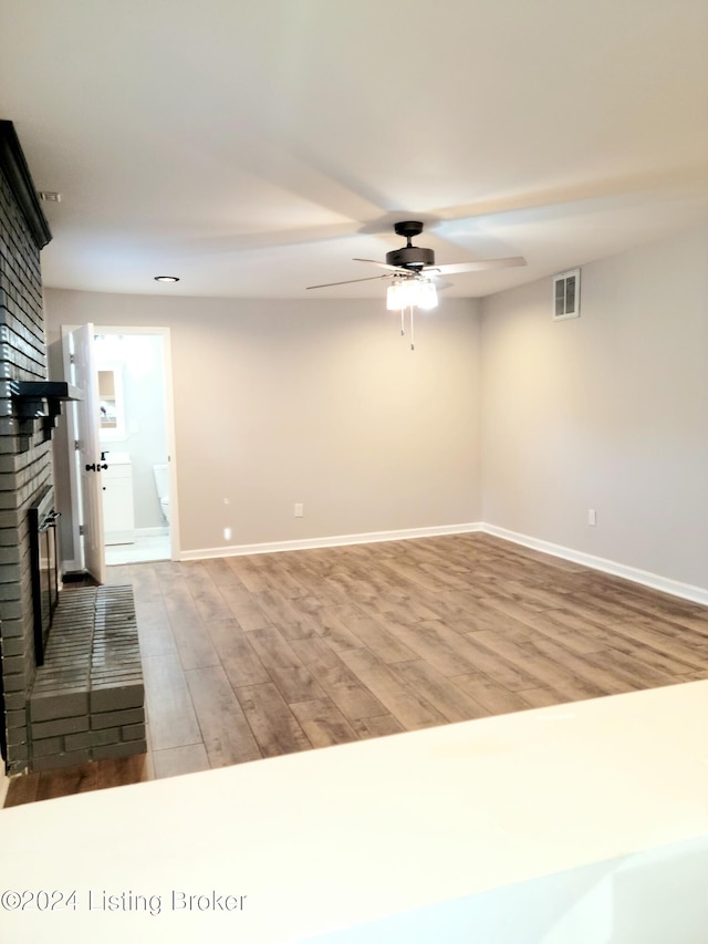 unfurnished room featuring a brick fireplace, wood-type flooring, and ceiling fan