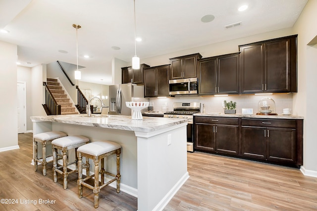 kitchen featuring light wood-type flooring, appliances with stainless steel finishes, decorative light fixtures, and an island with sink