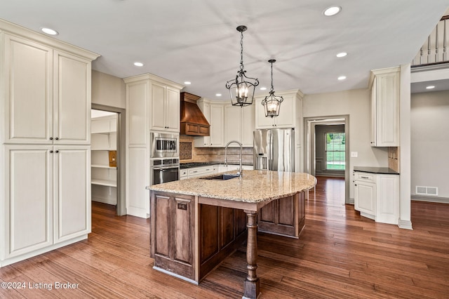 kitchen featuring stainless steel appliances, a center island with sink, sink, decorative light fixtures, and premium range hood