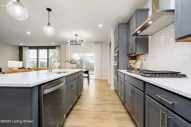 kitchen featuring wall chimney exhaust hood, sink, hanging light fixtures, a center island with sink, and appliances with stainless steel finishes