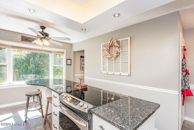 kitchen with ceiling fan, stainless steel range with electric cooktop, dark stone counters, a breakfast bar, and white cabinets