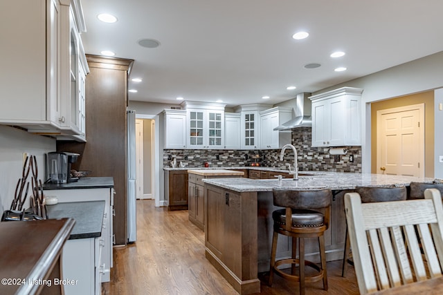 kitchen with hardwood / wood-style floors, light stone counters, a kitchen island with sink, and wall chimney exhaust hood