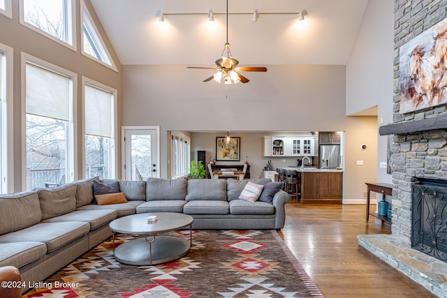 living room featuring high vaulted ceiling, a stone fireplace, rail lighting, ceiling fan, and light wood-type flooring