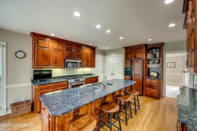 kitchen featuring dark stone counters, a center island with sink, sink, light wood-type flooring, and stainless steel appliances