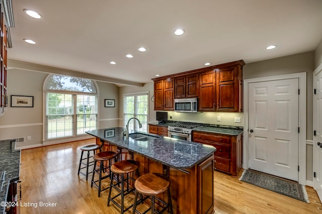 kitchen with sink, dark stone counters, a center island with sink, appliances with stainless steel finishes, and light wood-type flooring