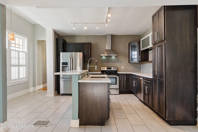 kitchen featuring sink, stainless steel appliances, wall chimney range hood, a kitchen island with sink, and dark brown cabinets