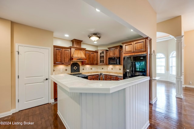 kitchen with dark wood-type flooring, black appliances, a center island with sink, decorative backsplash, and decorative columns