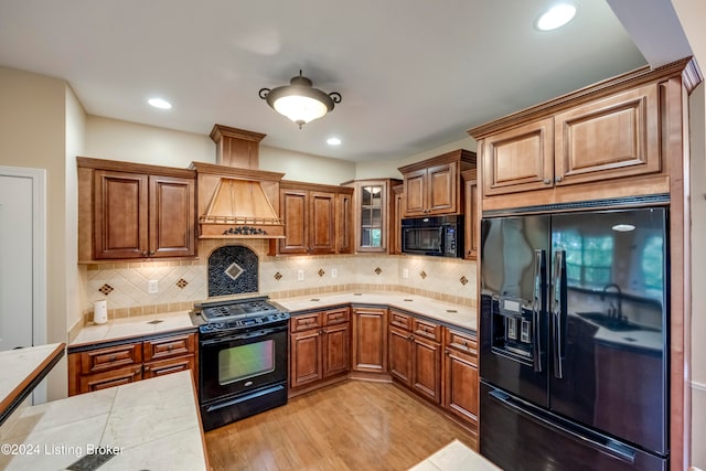 kitchen with tasteful backsplash, premium range hood, black appliances, light hardwood / wood-style flooring, and tile counters