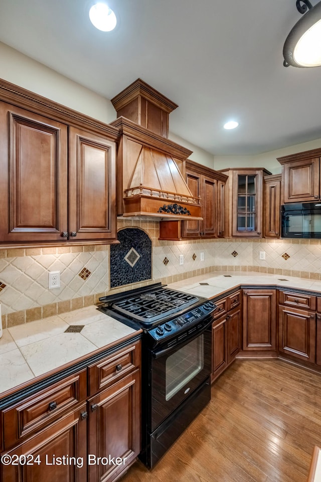 kitchen featuring black appliances, decorative backsplash, premium range hood, and light hardwood / wood-style flooring