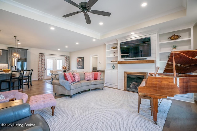 living room featuring a raised ceiling, built in features, ceiling fan, a fireplace, and light hardwood / wood-style floors