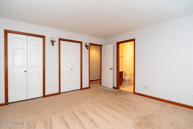 unfurnished bedroom with a textured ceiling, ensuite bathroom, light colored carpet, and two closets