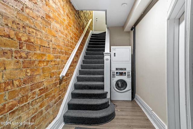 stairway with wood-type flooring, stacked washer and clothes dryer, and brick wall