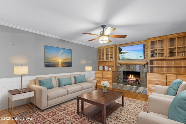 living room with a wainscoted wall, crown molding, a ceiling fan, a high end fireplace, and wood finished floors