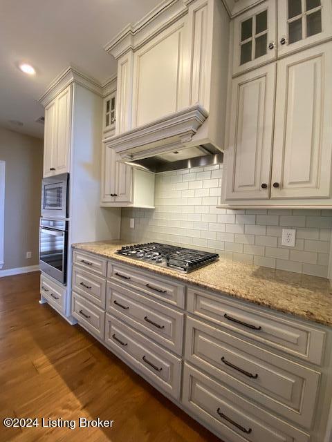 kitchen with backsplash, premium range hood, light stone counters, stainless steel appliances, and dark wood-type flooring