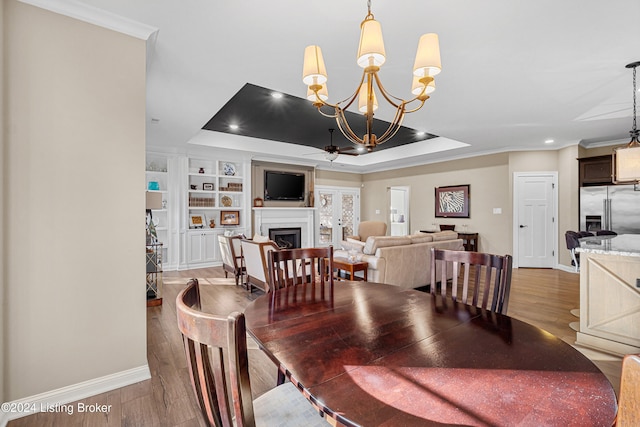 dining area with built in shelves, light hardwood / wood-style floors, a tray ceiling, ceiling fan with notable chandelier, and ornamental molding