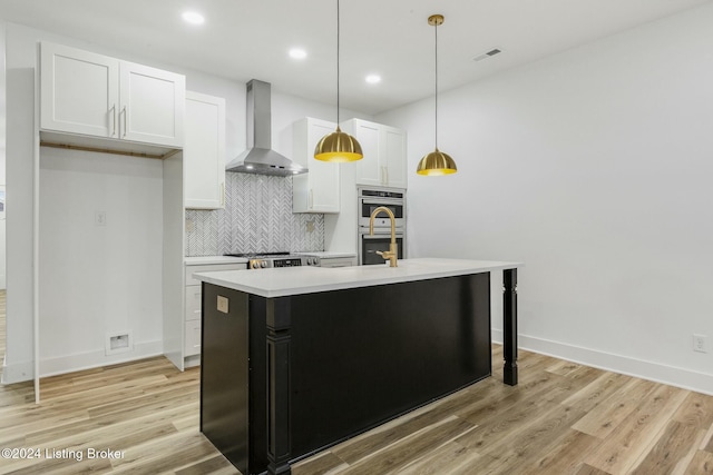 kitchen with a kitchen island with sink, wall chimney range hood, light hardwood / wood-style floors, white cabinetry, and hanging light fixtures