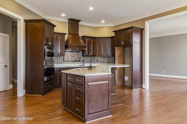 kitchen with premium range hood, a kitchen island with sink, oven, hardwood / wood-style flooring, and light stone countertops