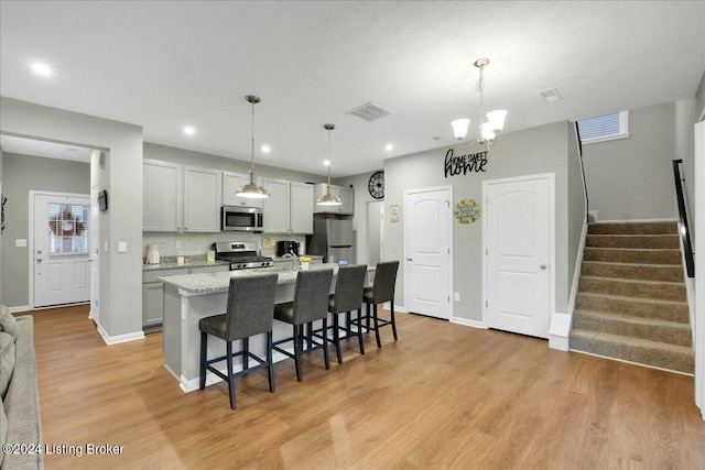 kitchen with gray cabinetry, light stone countertops, an island with sink, decorative light fixtures, and stainless steel appliances