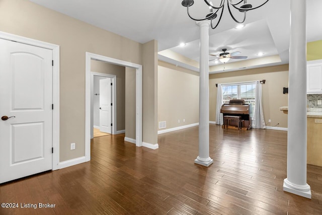 unfurnished living room with ceiling fan with notable chandelier, a tray ceiling, ornate columns, and dark wood-type flooring